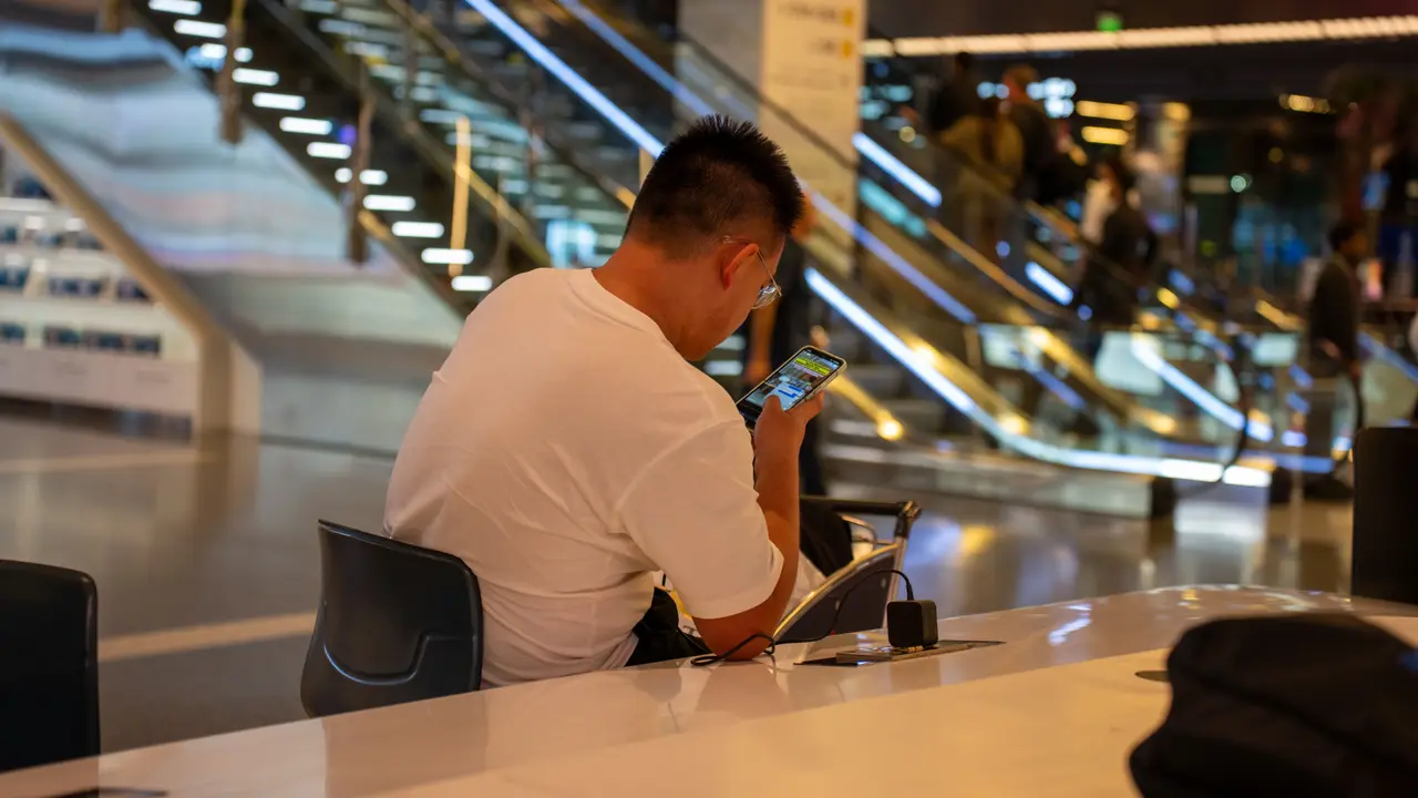 Una viajera mira el móvil en la terminal luminosa de un aeropuerto, con unas escaleras mecánicas al fondo.