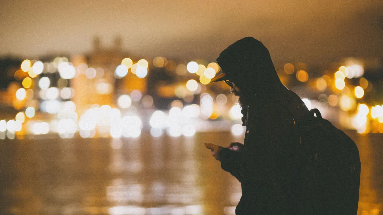 La silhouette d’un homme penché sur son téléphone, au bord d’un fleuve qui traverse la ville, la nuit.