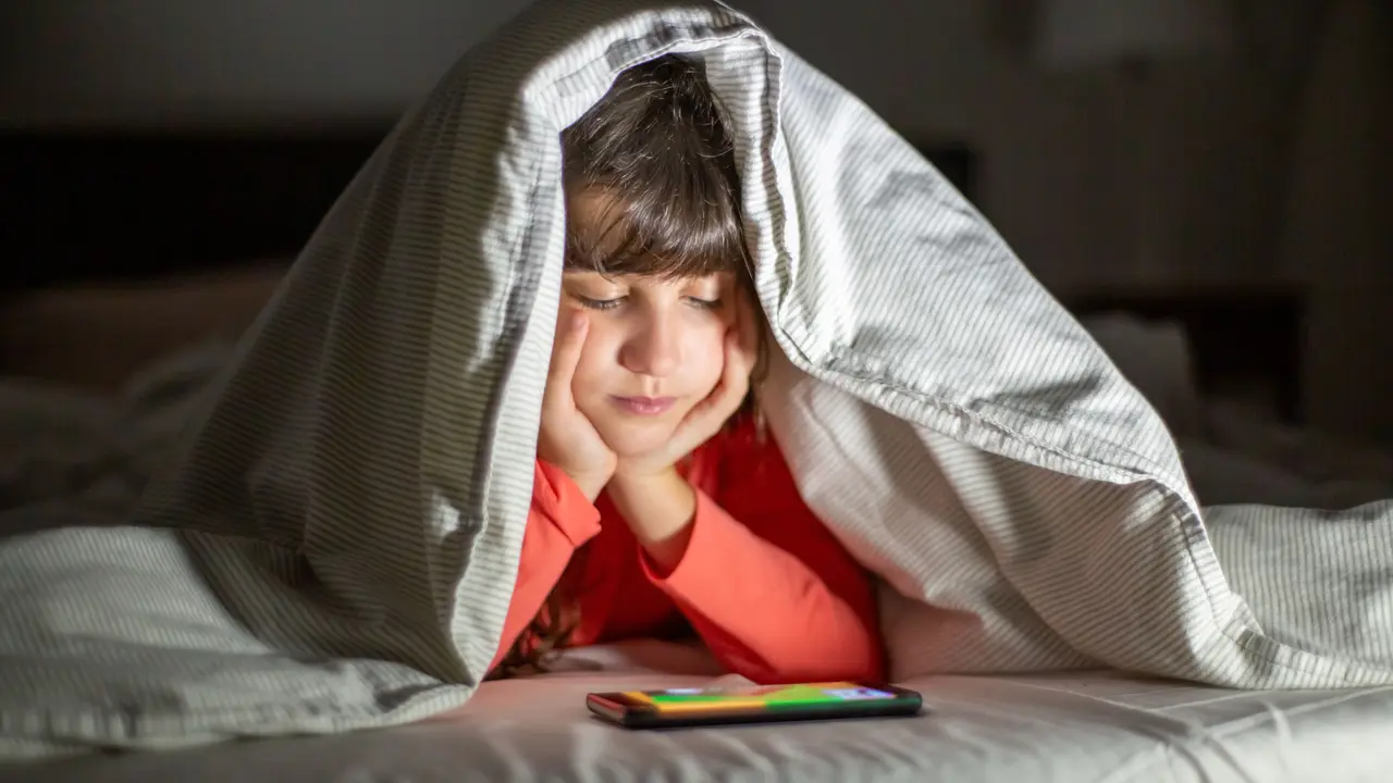 Un niño tapado con una manta viendo algo en la pantalla de un móvil.