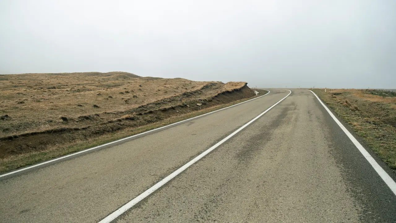 Eine einzelne Straße zieht sich durch eine leere, baumlose Landschaft unter dichter Wolkendecke.