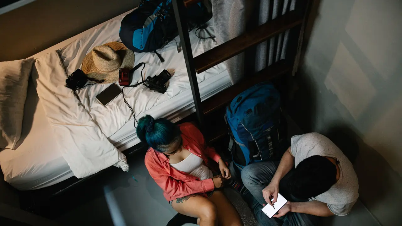 Una pareja con mochilas mira juntos la pantalla de un móvil en la habitación de un albergue.