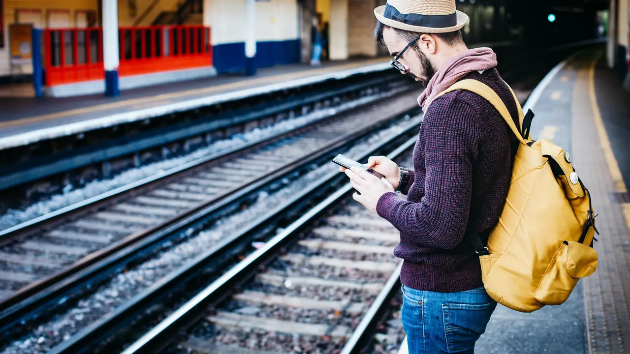 Ein Mann mit Rucksack schaut auf einem Bahnsteig auf sein Handy.