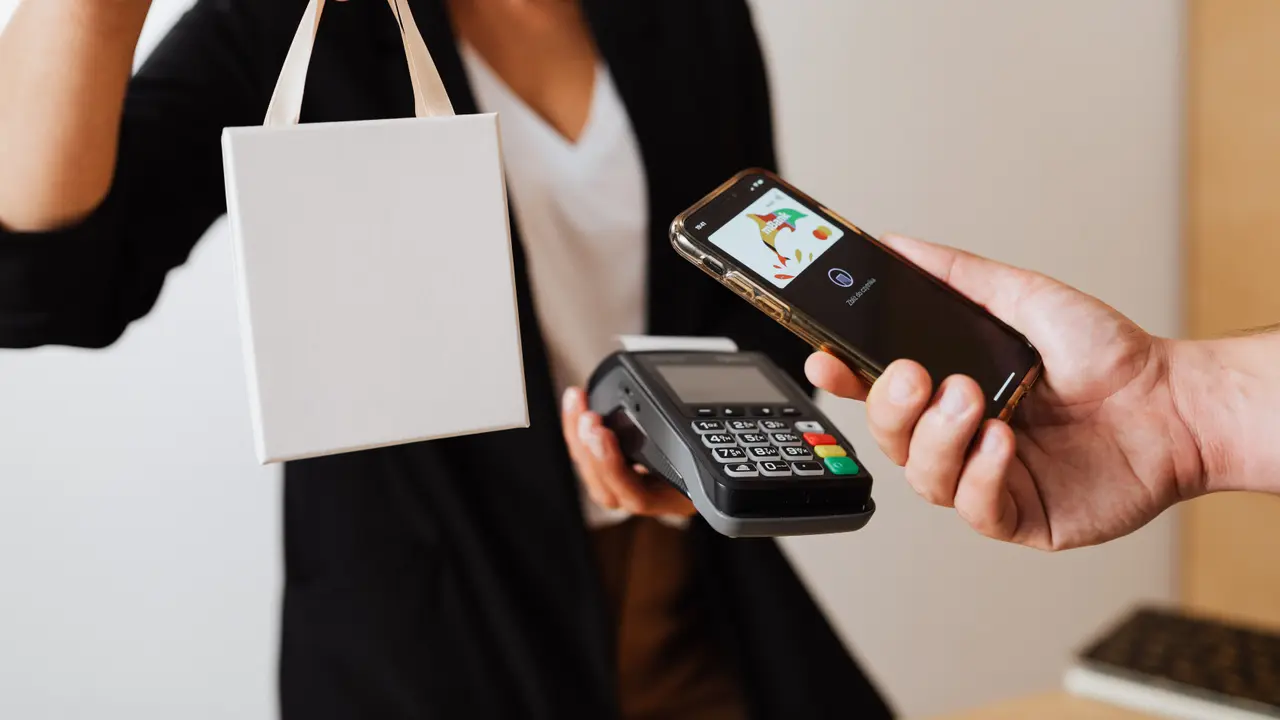 Bir el telefonu temassız kart terminaline tutarken tezgâhın arkasındaki satış görevlisi poşeti uzatıyor.