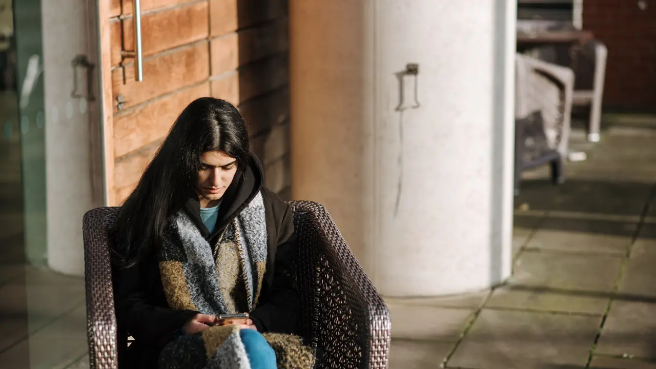 Una mujer abrigada, sentada en la terraza de un café, mira la pantalla del móvil que sostiene con las dos manos.