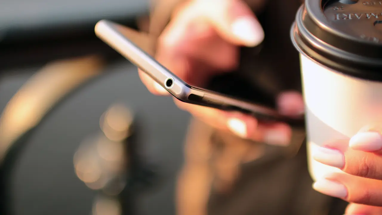 A close-up of a hand holding a phone and a coffee cup outdoors at sunset.