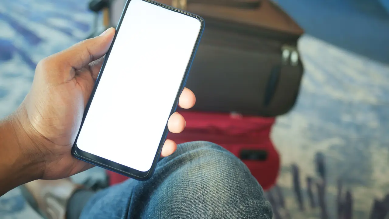 Eine Reisende steht mit dem Koffer in einer Flughafenhalle und blickt nach unten auf ihr Handy.