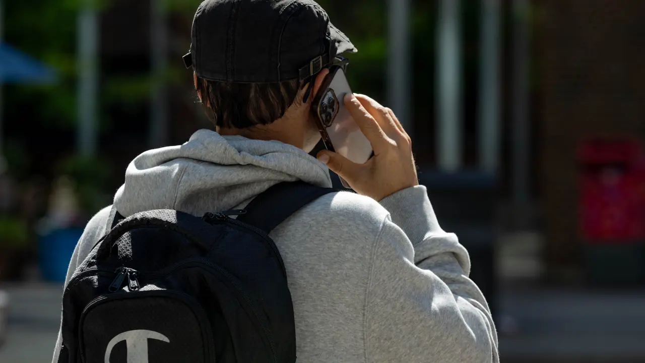 Un uomo con lo zaino, ripreso di spalle, parla al telefono all’aperto.