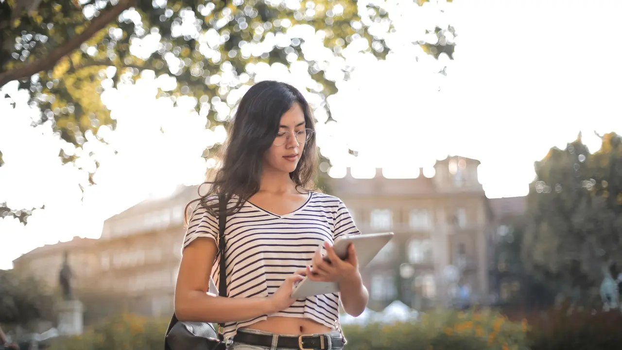 A young woman in a striped shirt using a tablet outdoors in a sunlit square, a historic building behind her.