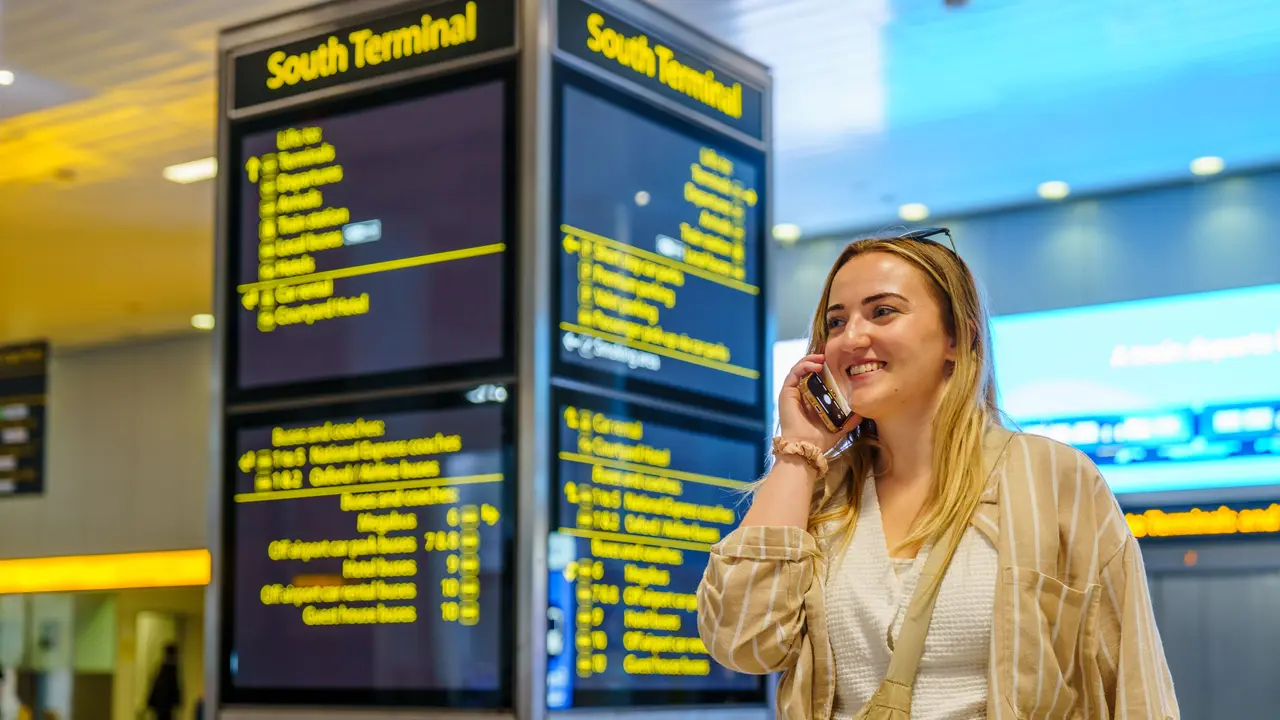 A woman smiling on a phone call in an airport terminal, departure boards behind her.