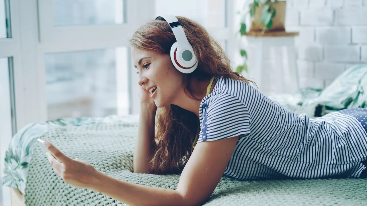 Eine junge Frau liegt mit Kopfhörern auf einem Bett und schaut auf ihr Handy.