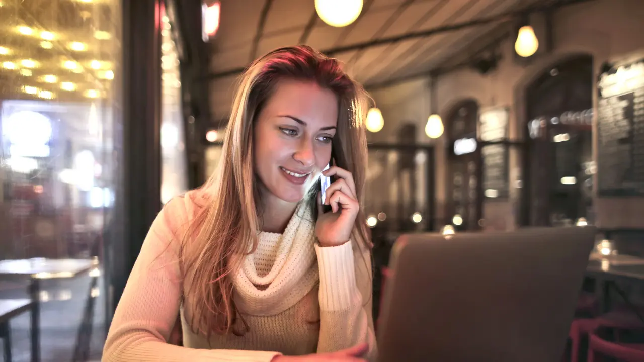 Eine Frau sitzt in einem Café und telefoniert mit ihrem Handy, vor ihr steht ein aufgeklappter Laptop.